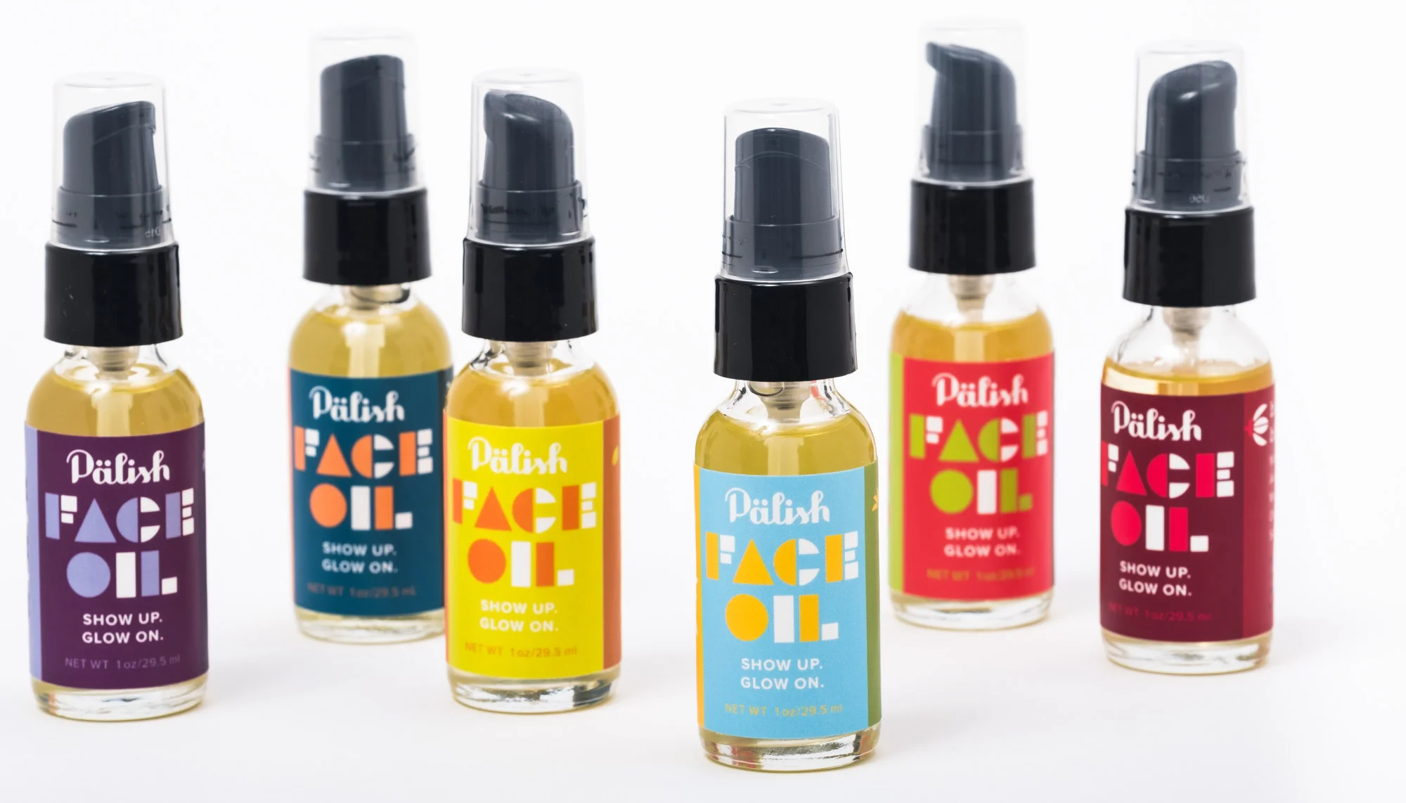 Six small bottles of face oil with colorful labels and black spray caps, arranged in a row.