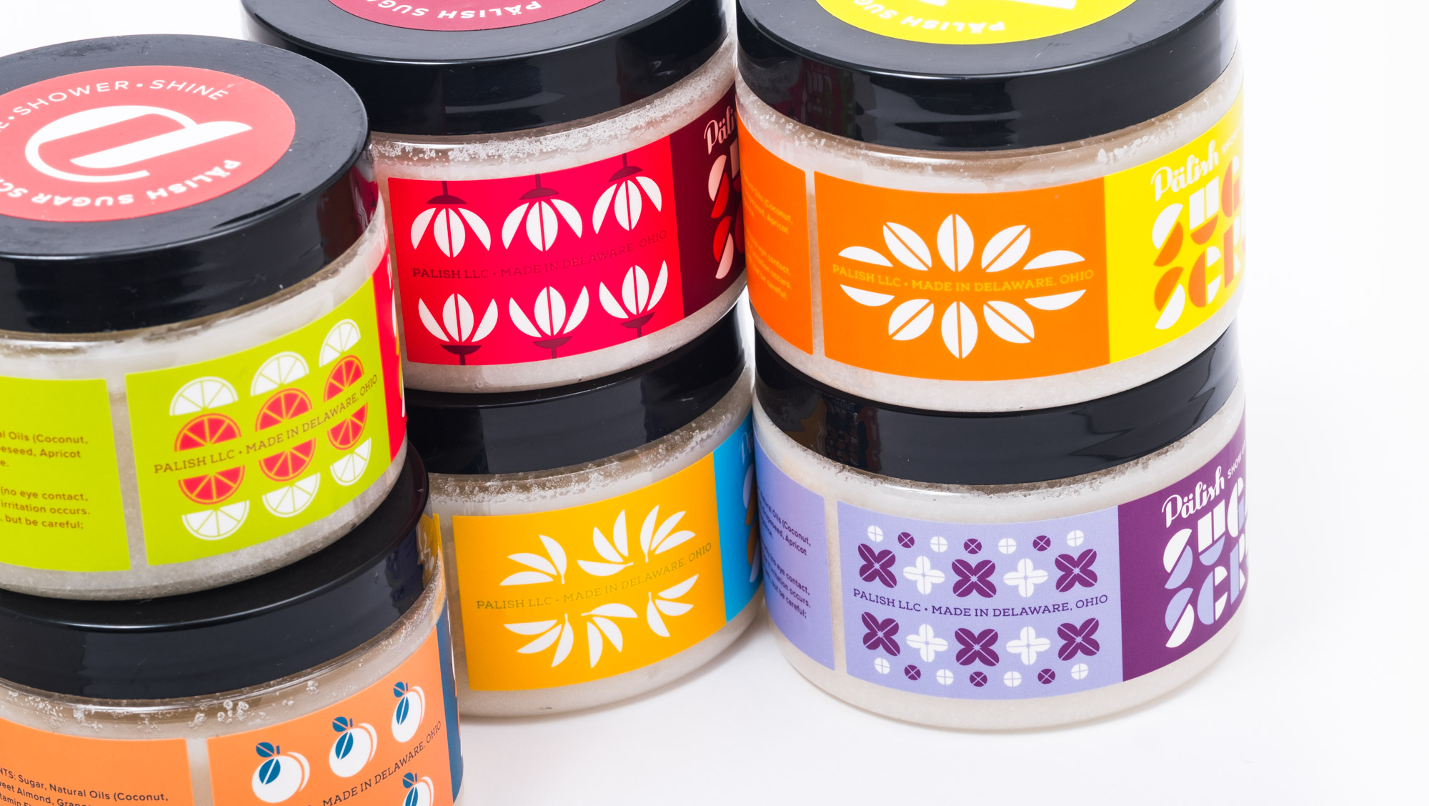 Stacked jars of body scrub with colorful labels in yellow, orange, purple, red, and blue, featuring citrus and floral designs, on a white background.