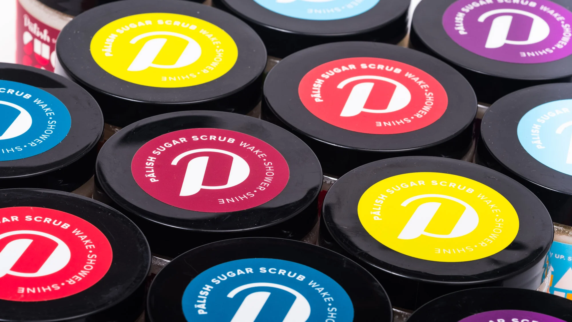 Close-up of colorful jars of sugar scrub with black lids, featuring circular labels in yellow, red, purple, and blue with white text and logos.