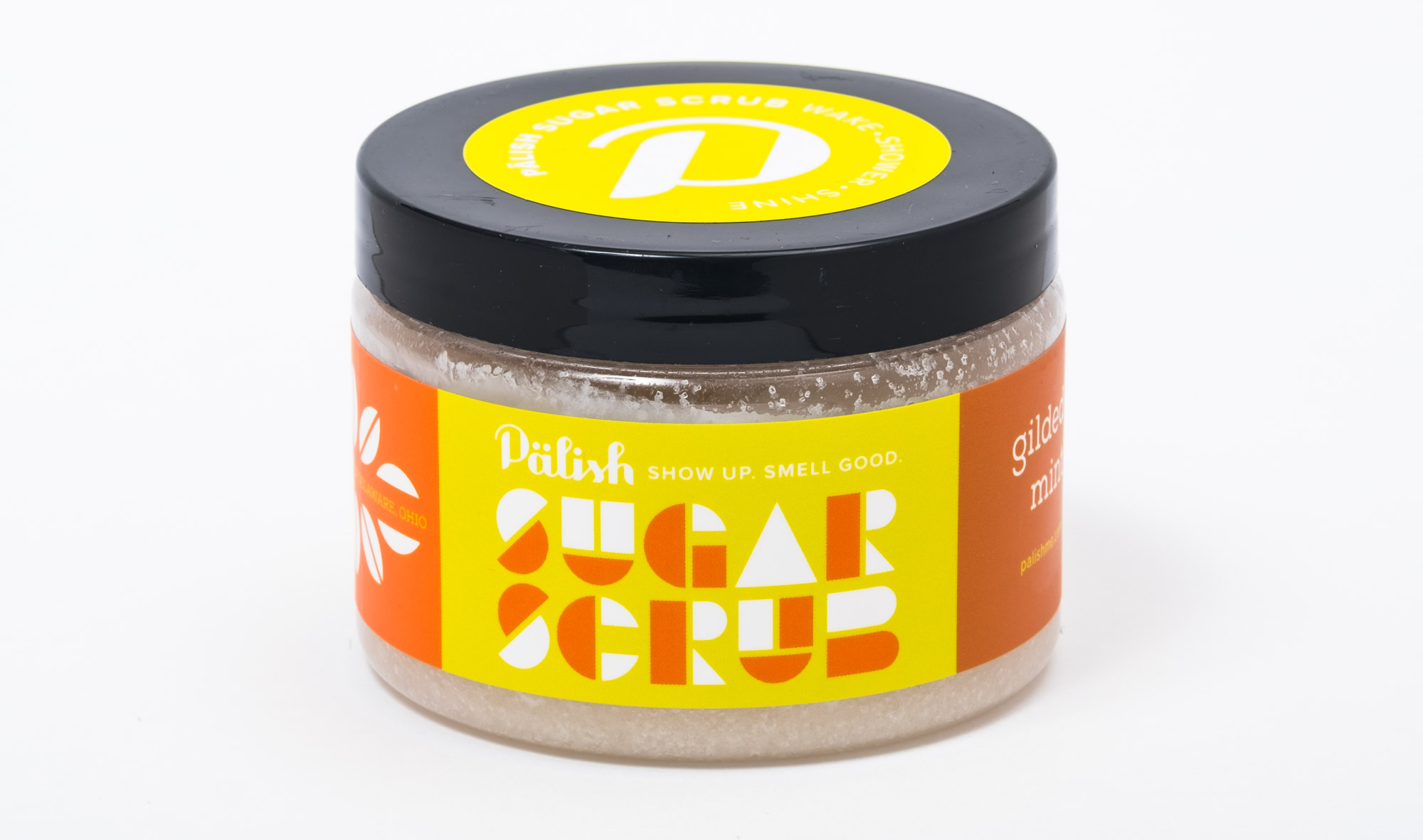 A jar of pink sugar scrub with a black lid and a yellow and orange label that says 'PAllsh. Show up. Smell good. Sugar Sugar.'