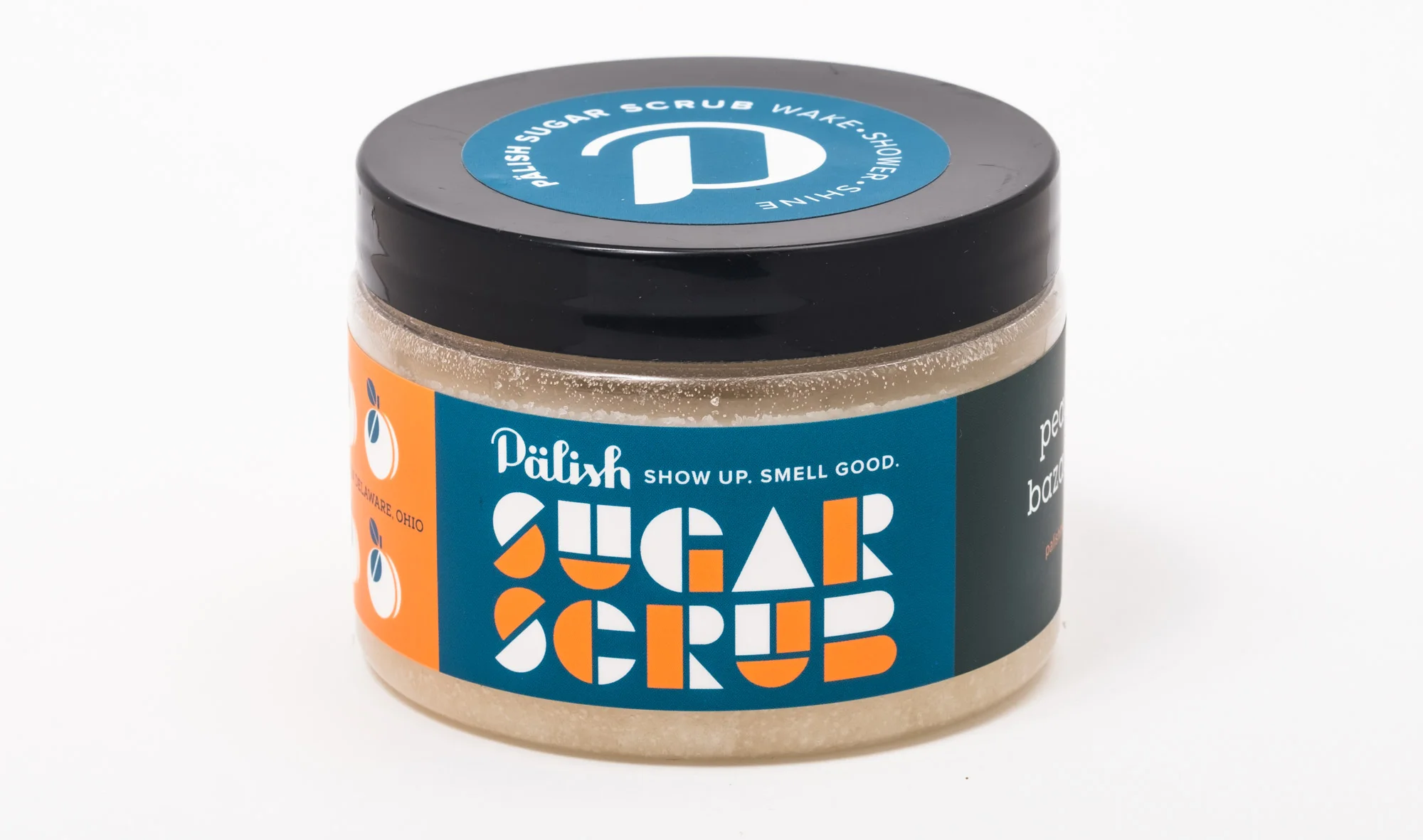 A jar of Pālish Sugar Scrub with blue, orange, and white packaging. The label says 'SHOW UP. SMELL GOOD.' and 'Sugar Scrub.'