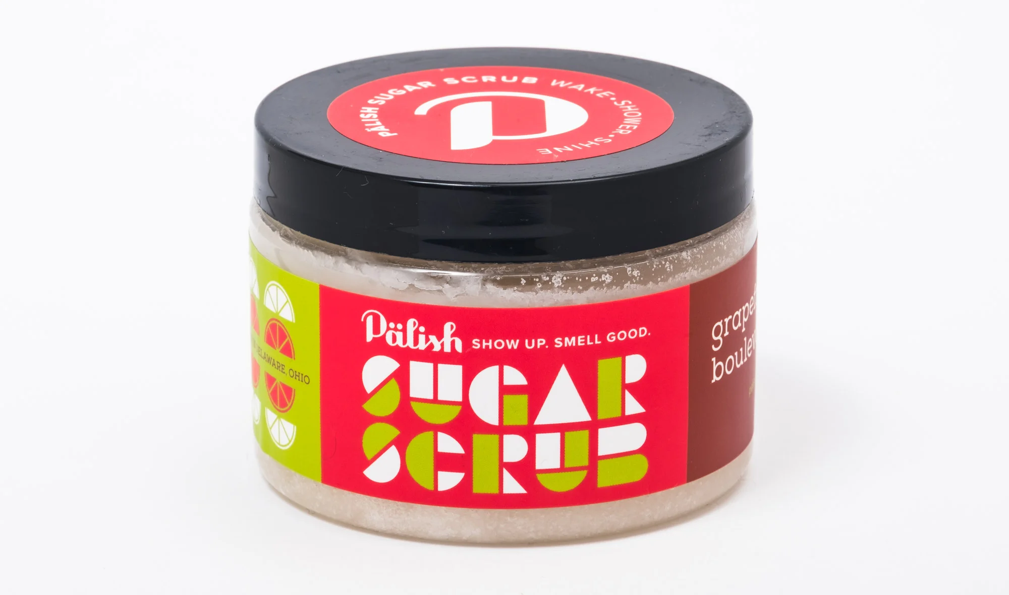 A small jar of pink grapefruit sugar scrub with a black lid and red label that reads 'Pälish Sugar Scrub' and 'show up. smell good.'