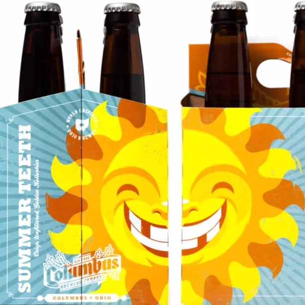 Six brown glass bottles in a cardboard carrier with a smiling sun face graphic, indicating summer refreshment from Columbus Brewing Company in Ohio.