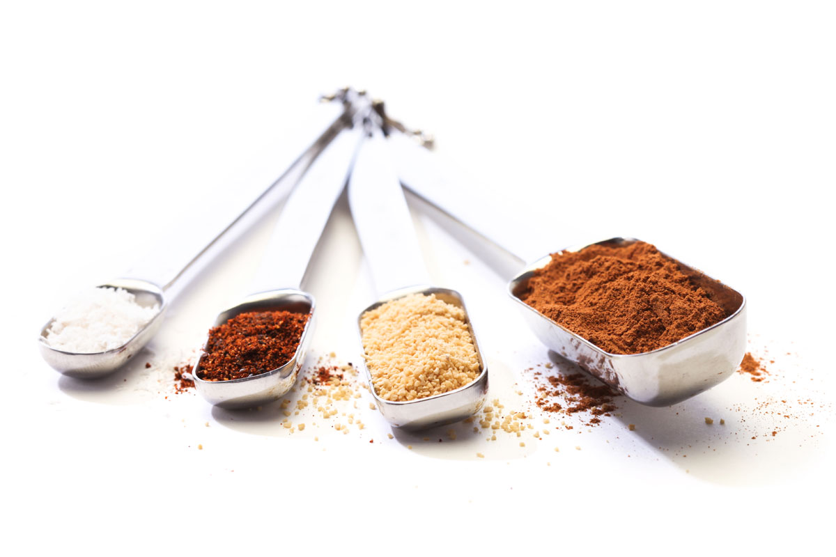 Four metal scoops filled with different spices, arranged in a fan shape, on a white background.