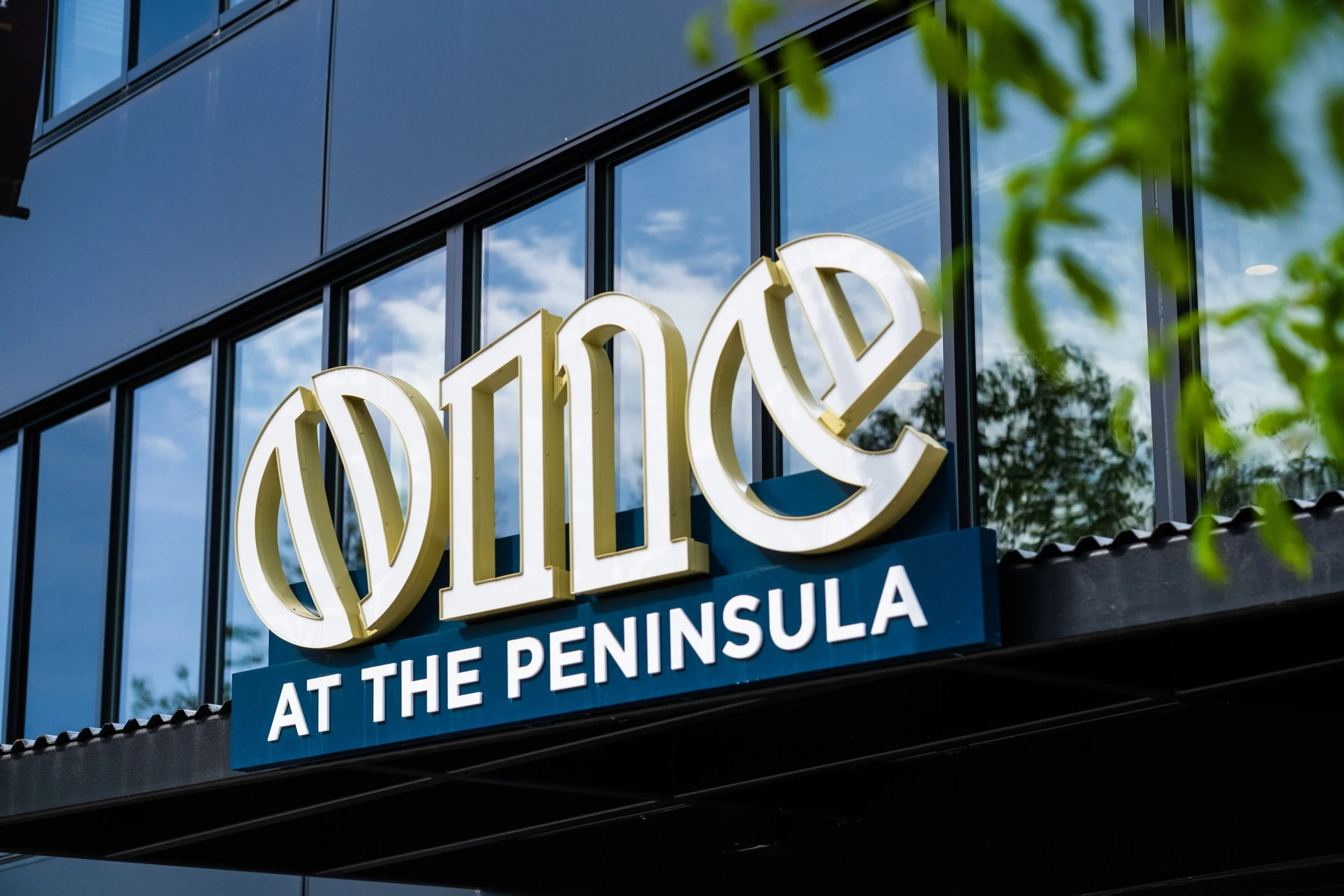 Sign for Wild at the Peninsula restaurant, featuring large white and gold letters and a blue background, reflected in the glass windows of a building, with green leaves partially visible.