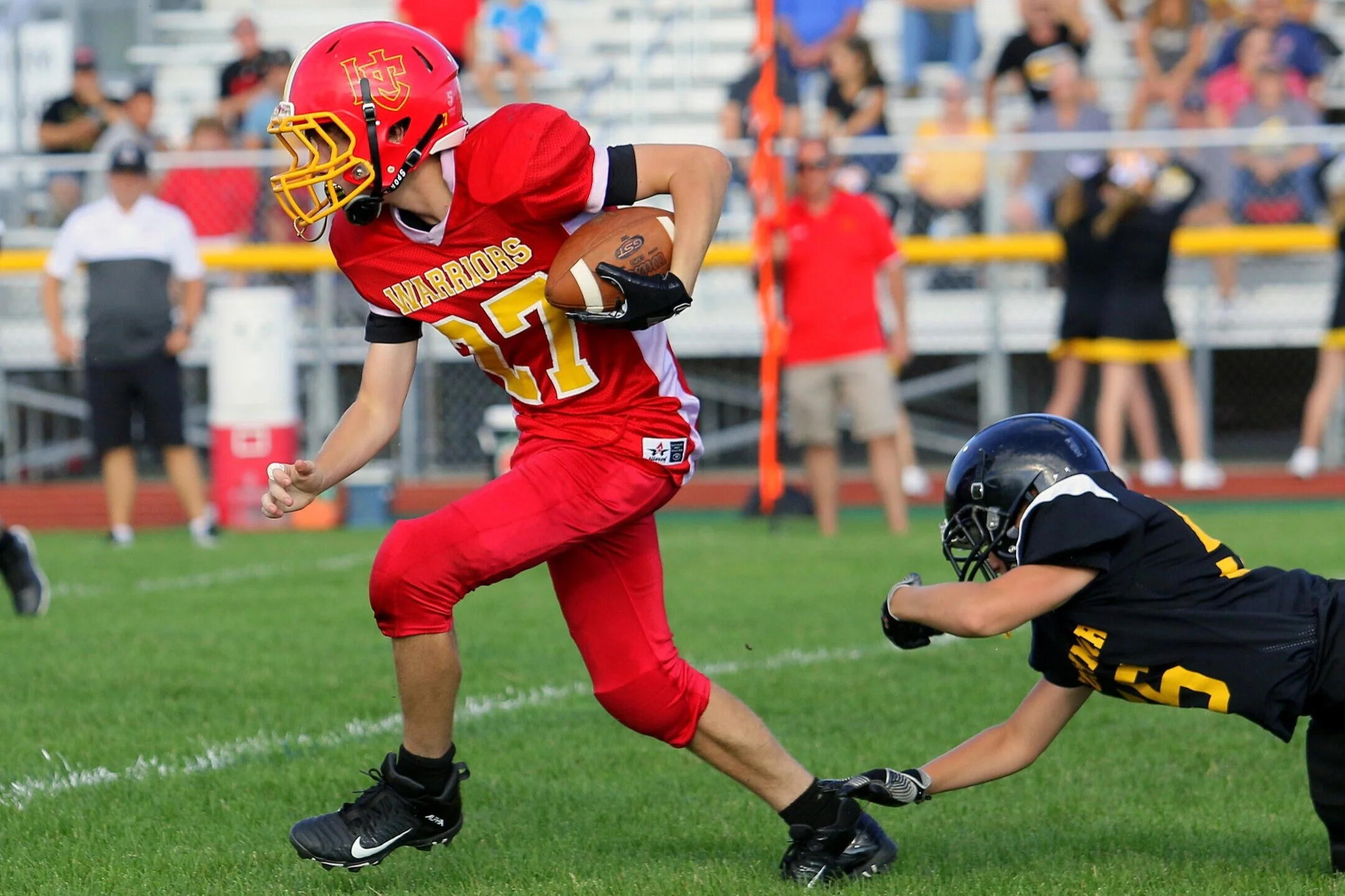 A football player wearing a red and yellow uniform with the number 21 is running on the field while holding a football. An opposing player in a black uniform and helmet is diving to tackle him.