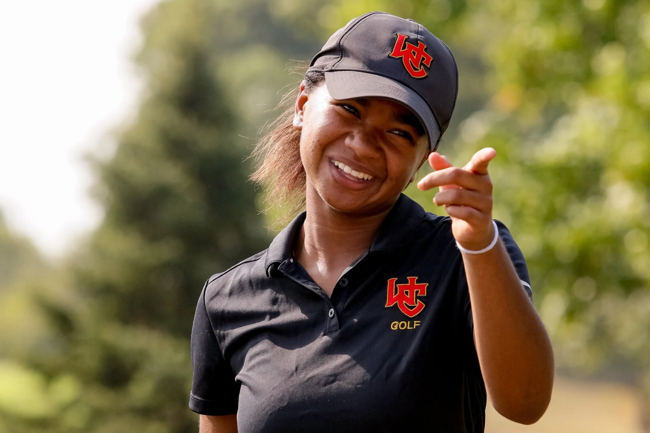 Young woman smiling and pointing outdoors, wearing a black golf shirt and cap with a red and yellow logo.