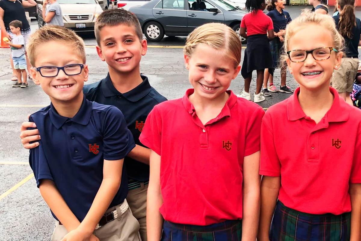 Four children, two boys and two girls, standing together outside on what appears to be a school event or gathering. They are smiling and wearing polo shirts with a school logo, in red and navy blue, and plaid skirts or shorts.