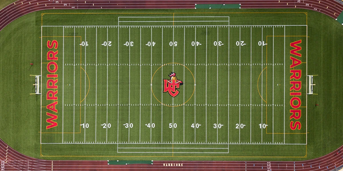 An aerial view of a football field with the word 'WARRIORS' written in red on both sides of the field, and the center logo depicting a warrior helmet with a red 'W'. The field is surrounded by a running track and has white and yellow yard lines marki