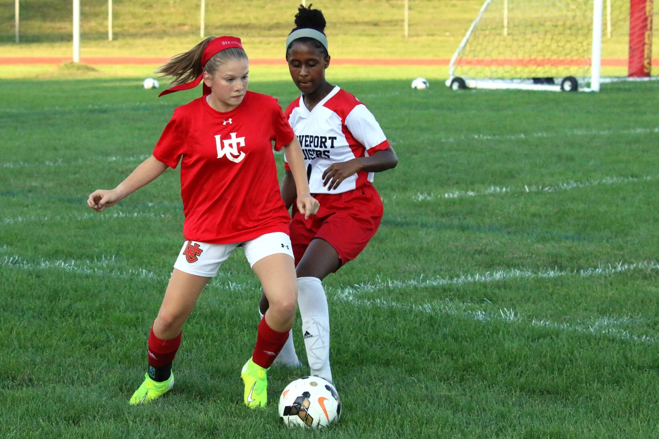 Two young female soccer players competing for control of a soccer ball on a grassy field, with goal net in the background.