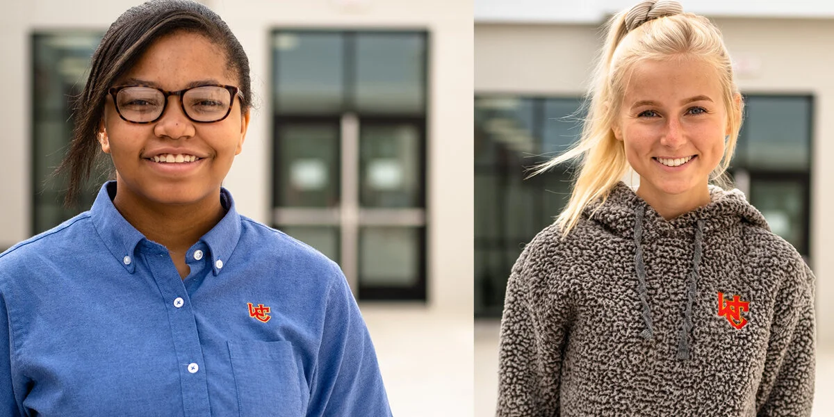 Two young women standing outside in front of a modern building, smiling at the camera. The woman on the left has dark hair, glasses, and wears a blue button-up shirt. The woman on the right has blonde hair tied back and wears a fuzzy gray hoodie. Bot