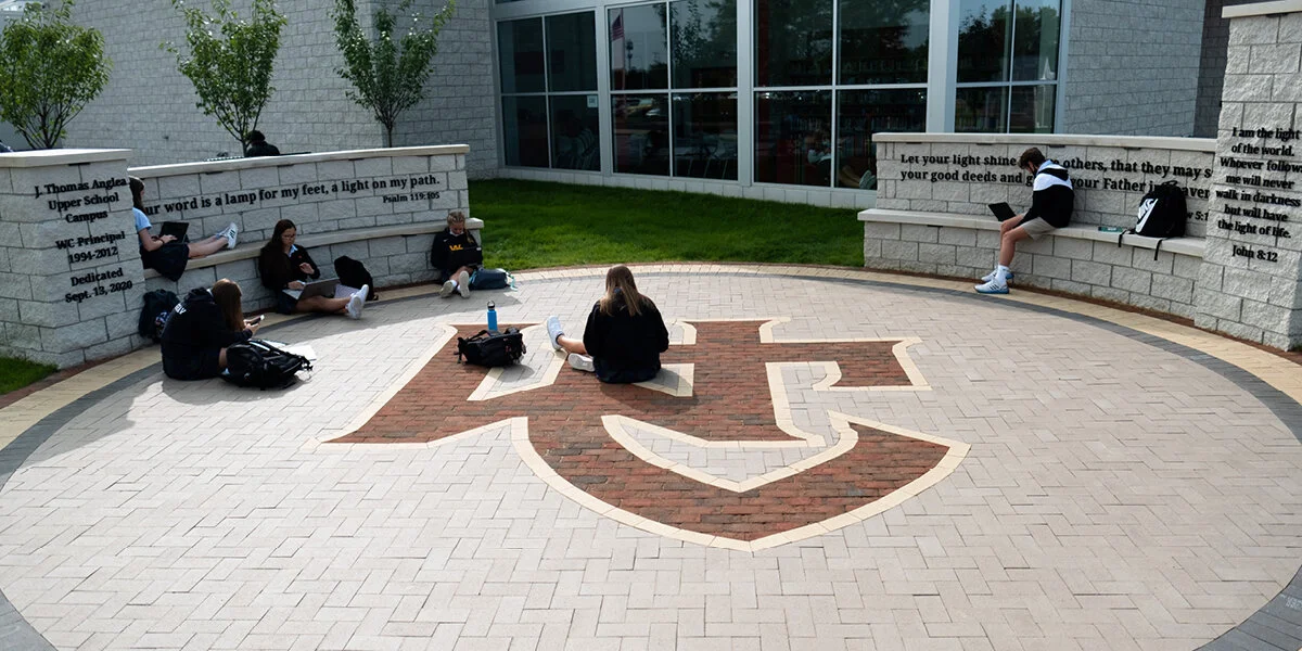 Students sitting on a circular brick-patterned area with a shield emblem, outside a modern building with large glass windows. Two students are on a curved bench with inscribed quotes, and others are seated on the ground with backpacks.