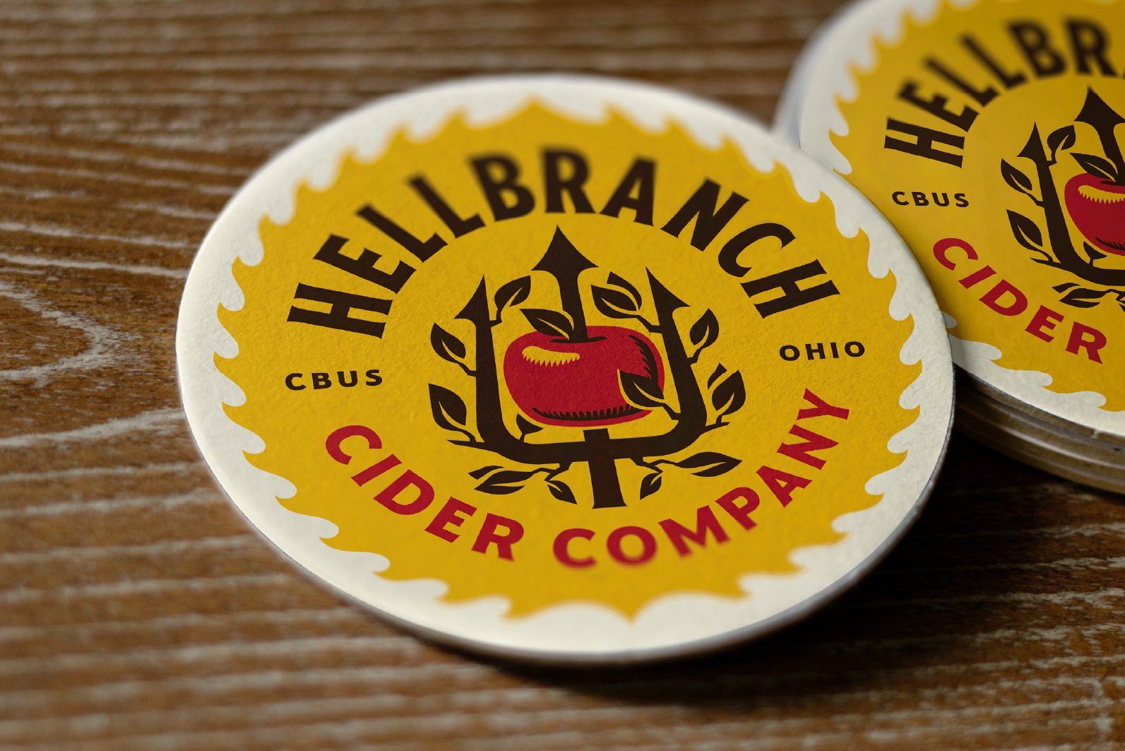 A sticker with a yellow background and black and red text that reads 'Hellebach Cider Company,' featuring a red apple surrounded by black leaves and a black trident, on a wooden surface.