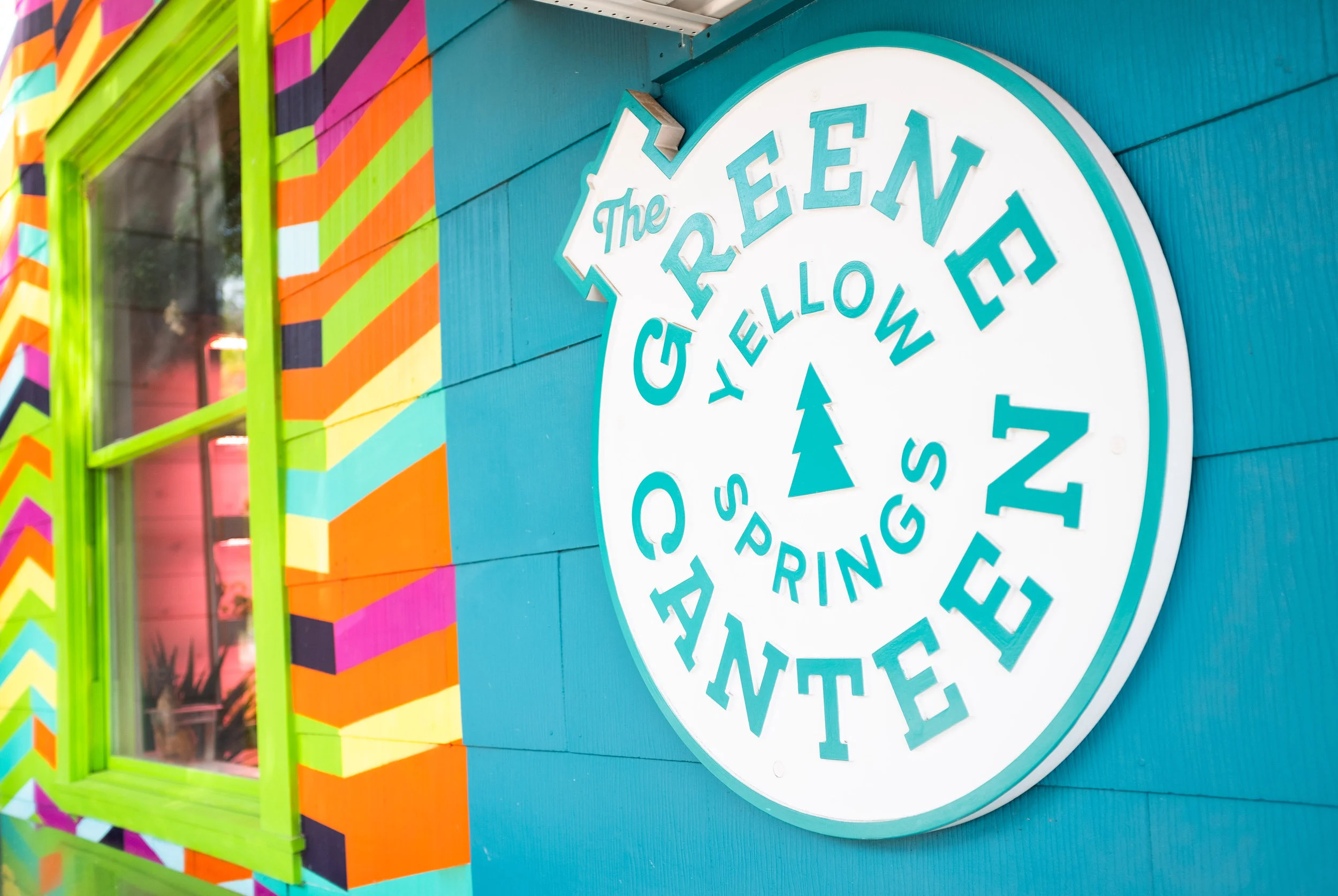 Colorful storefront with a round sign reading 'GIVE LOVE THE REE N'. The sign has a pine tree symbol at the center. The building features bright, multicolored zigzag patterns around a green-framed window.