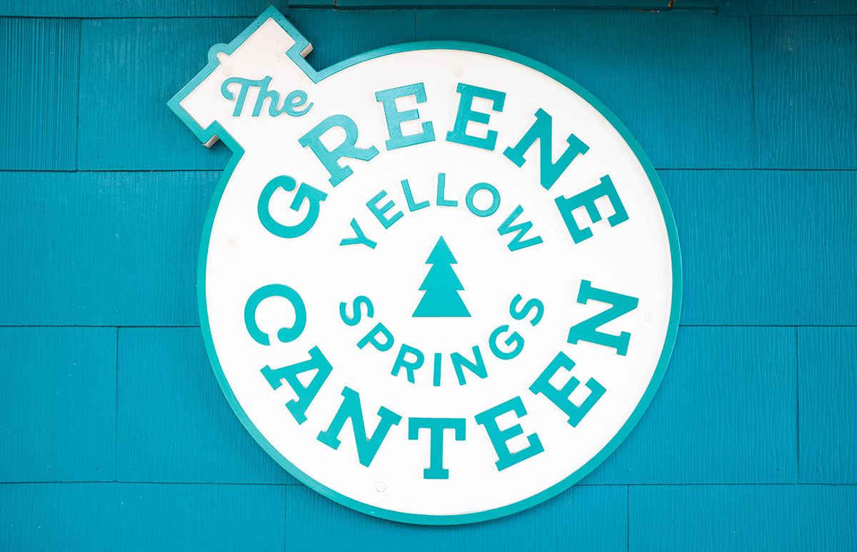 Sign for The Green Canteen with yellow springs in teal color, featuring a circular design with a pine tree graphic in the center, mounted on a teal wooden wall.