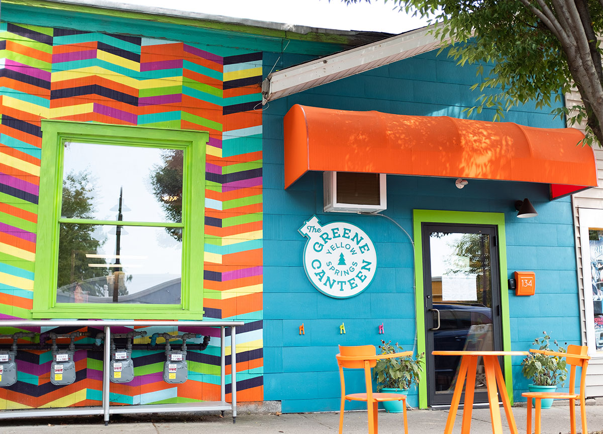 Colorful building exterior with a blue wall, a bright green window frame, and a multicolored striped wall section. There is an orange awning above a black door, and orange chairs with small plants on their tables outside.