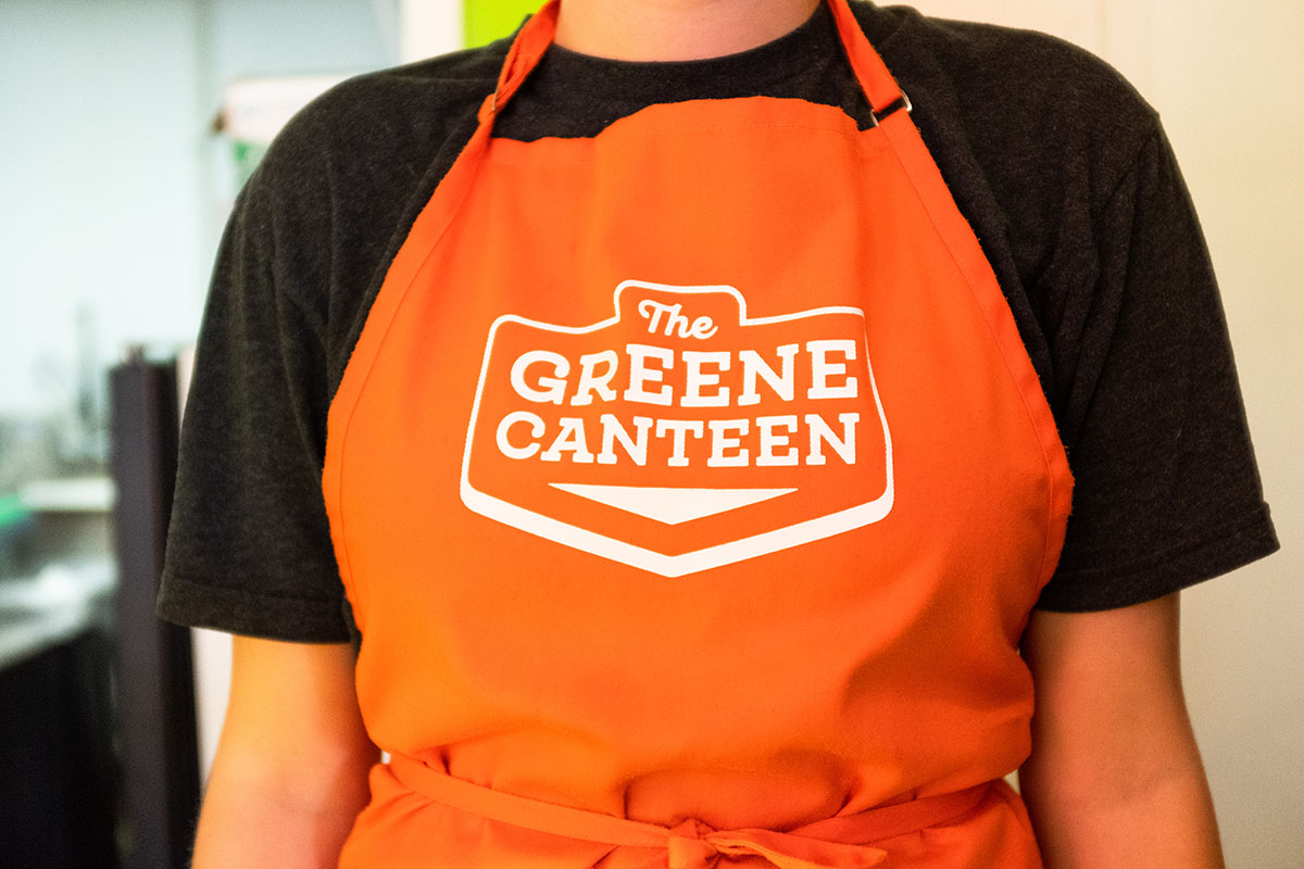 Person wearing an orange apron with the logo 'The Greene Canteen' printed on it, over a black t-shirt.
