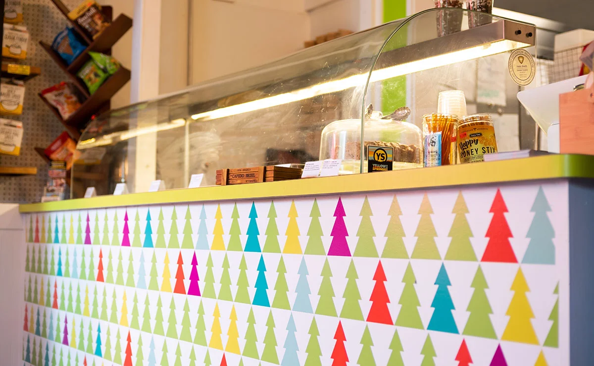 Candy shop counter with a glass display case, colorful triangle patterned front panel, and snacks on the shelves behind.