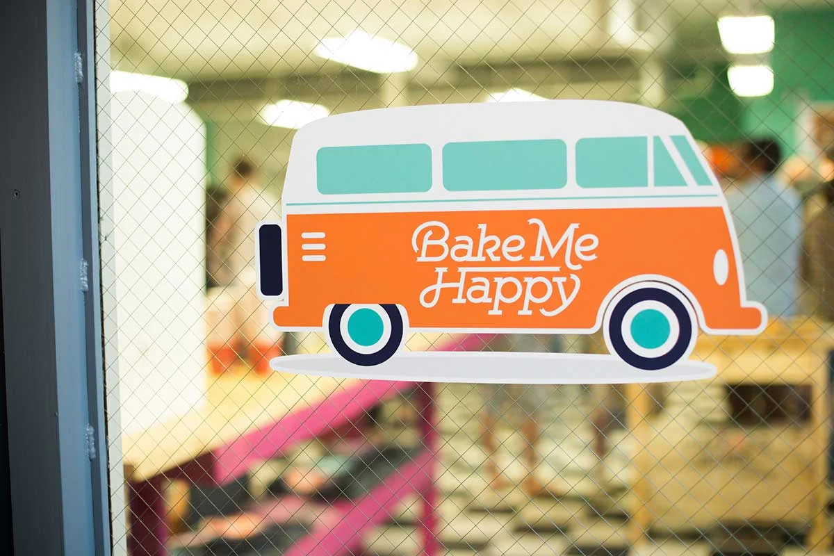 A sticker of an orange and white camper van with the words "Bake Me Happy" on it, attached to a glass door, with a blurred bakery interior in the background.