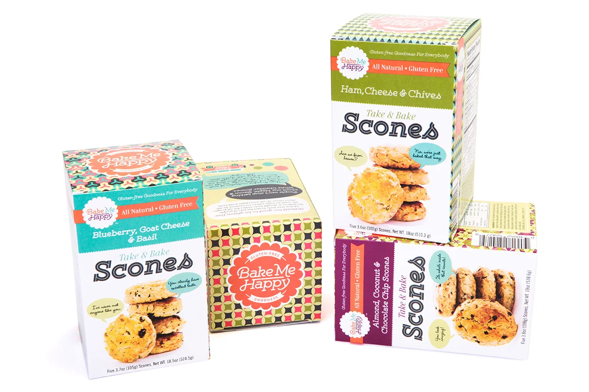 Three boxes of gluten-free Bake Me Happy scone mixes, including Blueberry Goat Cheese & Basil, Ham Cheese & Chives, and Almond & Chocolate Chip, with a decorative cube in the center.