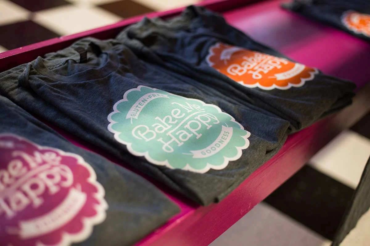 Folded dark gray T-shirts with a colorful badge design that says 'Bake Me Happy Goodness' displayed on a pink tray.