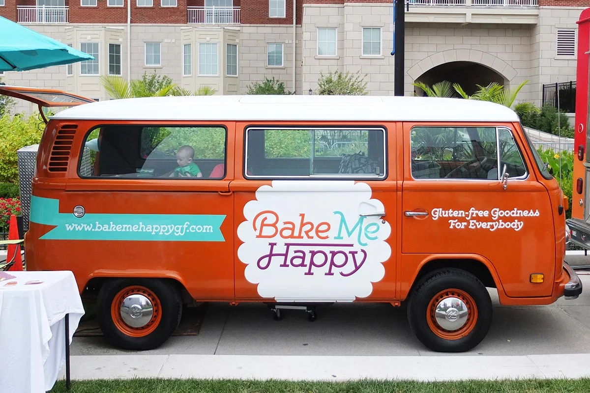 An orange and white van with signage for Bake Me Happy, a gluten-free bakery. The side of the van displays the website www.bakemehappygf.com and the slogan 'Gluten-free Goodness For Everybody.' The van is parked outside, with a child visible through 