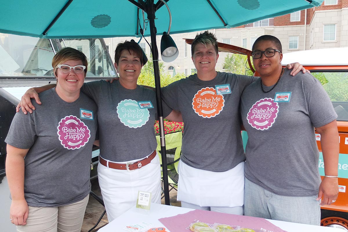 Four women standing side by side smiling at an outdoor event. They are wearing matching gray t-shirts with colorful 'Bake Me Happy' badges on the front. One woman has her arm around another, underneath a large teal umbrella, with a food truck or vend