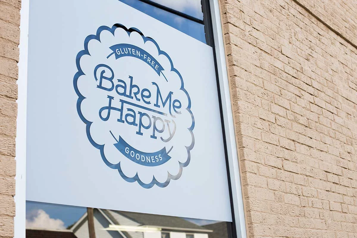 Sign for a gluten-free bakery named Bake Me Happy with the phrases 'Gluten-Free' and 'Goodness' on a blue and white logo on the window of a brick building.