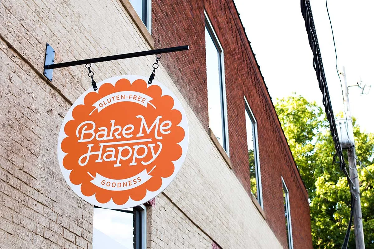 A bakery sign hanging outside on a brick wall, reading "Bake Me Happy," with the words "Gluten-Free" and "Goodness" included in the design, and a background of green trees and a blue sky.