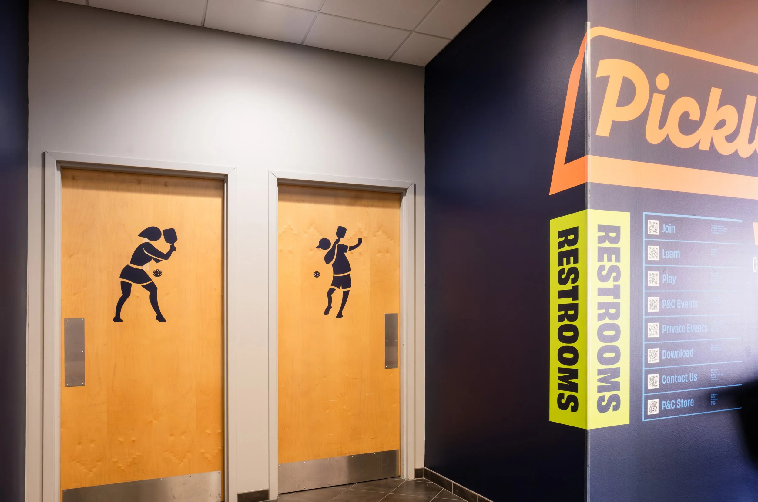 Two elevator doors with simple black silhouette illustrations of children playing. The left door shows a girl holding a racket, and the right door shows a girl holding a tennis ball and racket.