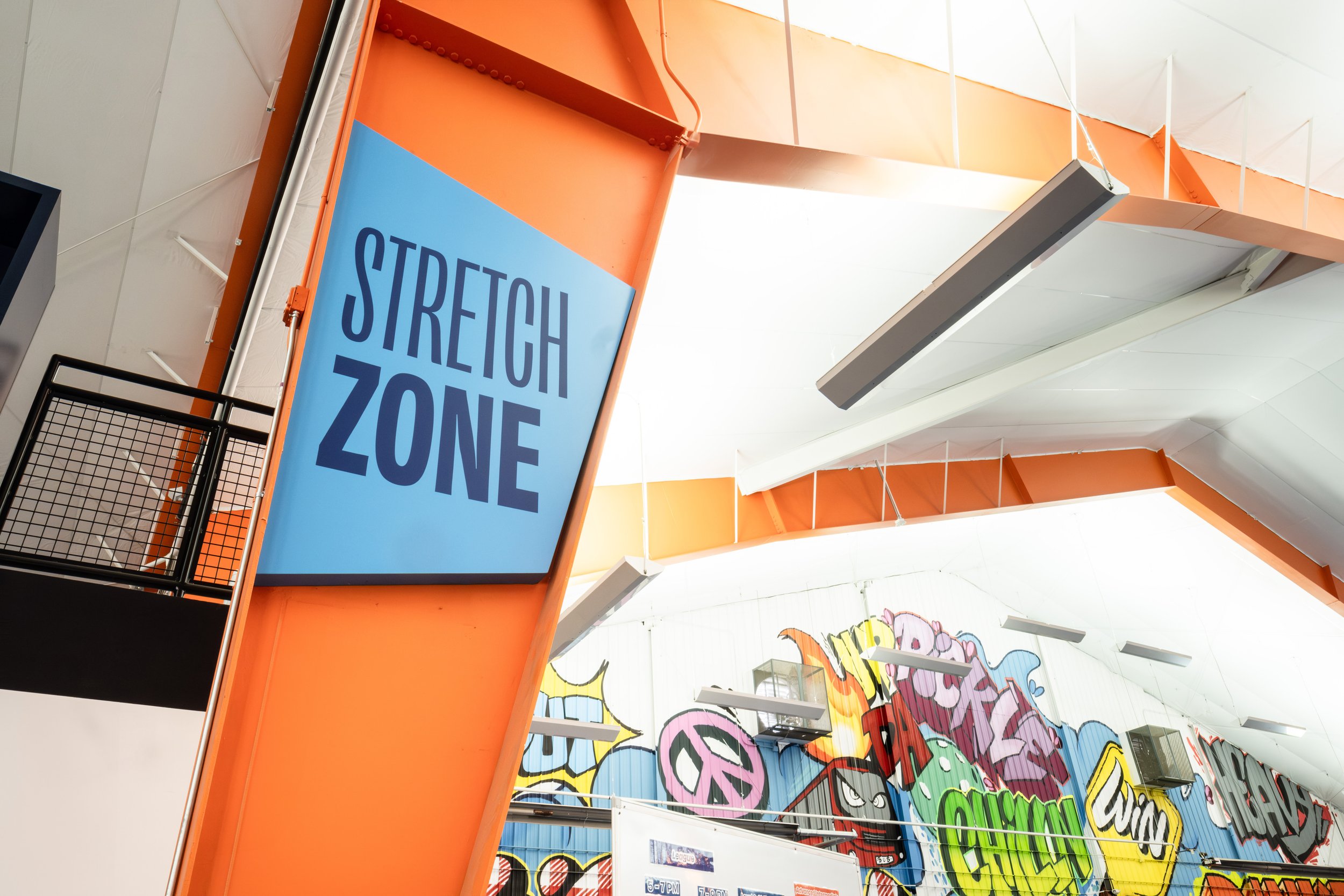 Bright orange sign with blue and dark blue text reading 'STREET ZONE' hanging from the ceiling of a colorful graffiti-covered indoor space.