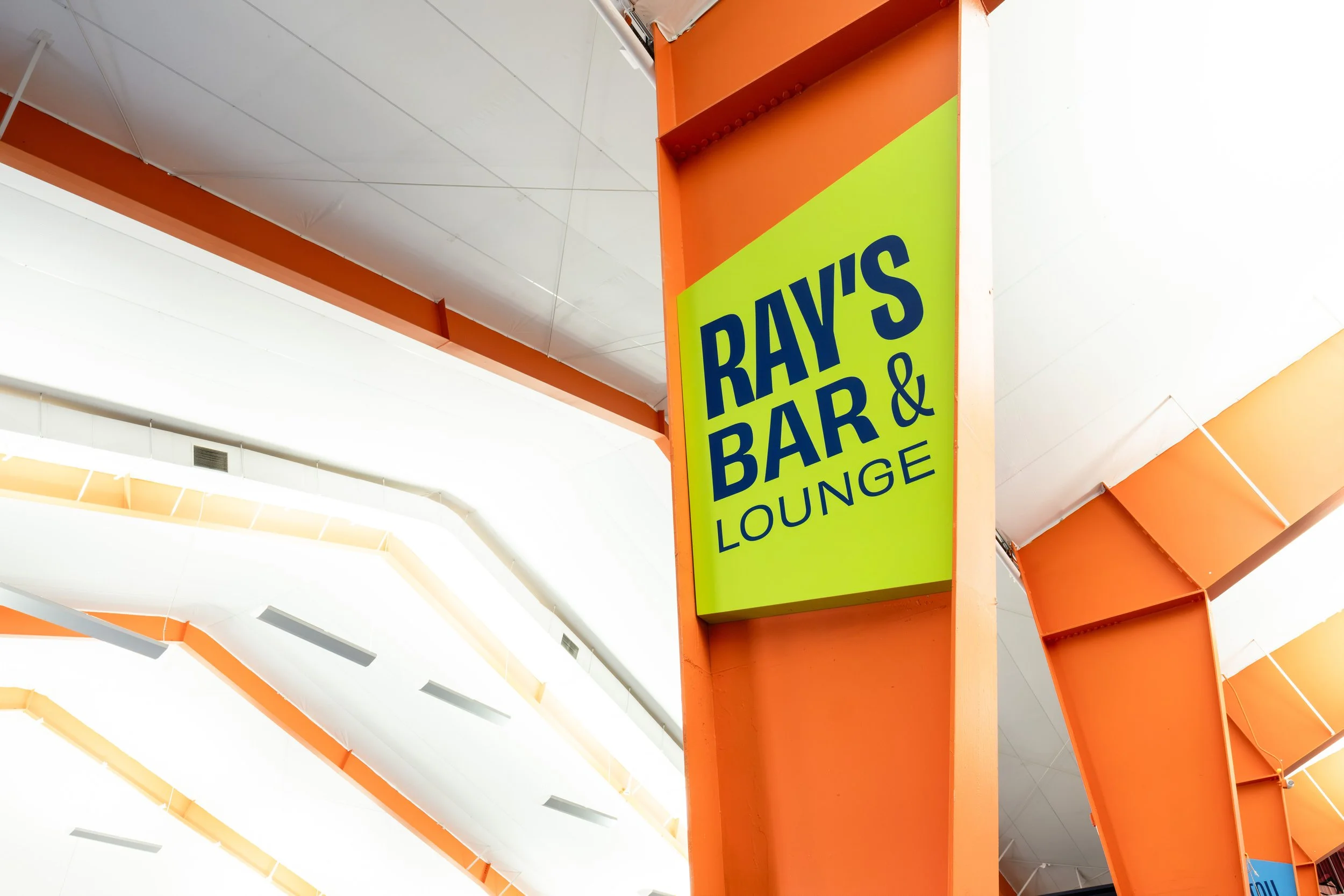 A bright green sign with blue text that reads 'Ray's Bar & Lounge' is attached to an orange structural element inside a building, with a white ceiling and ceiling fans visible in the background.