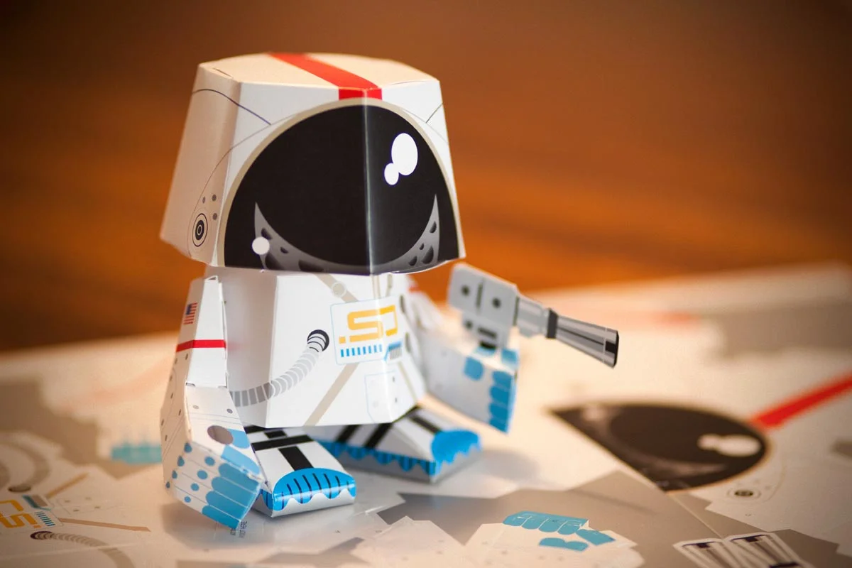 A paper toy robot in an astronaut suit holding a small gun, surrounded by paper cutouts on a table.