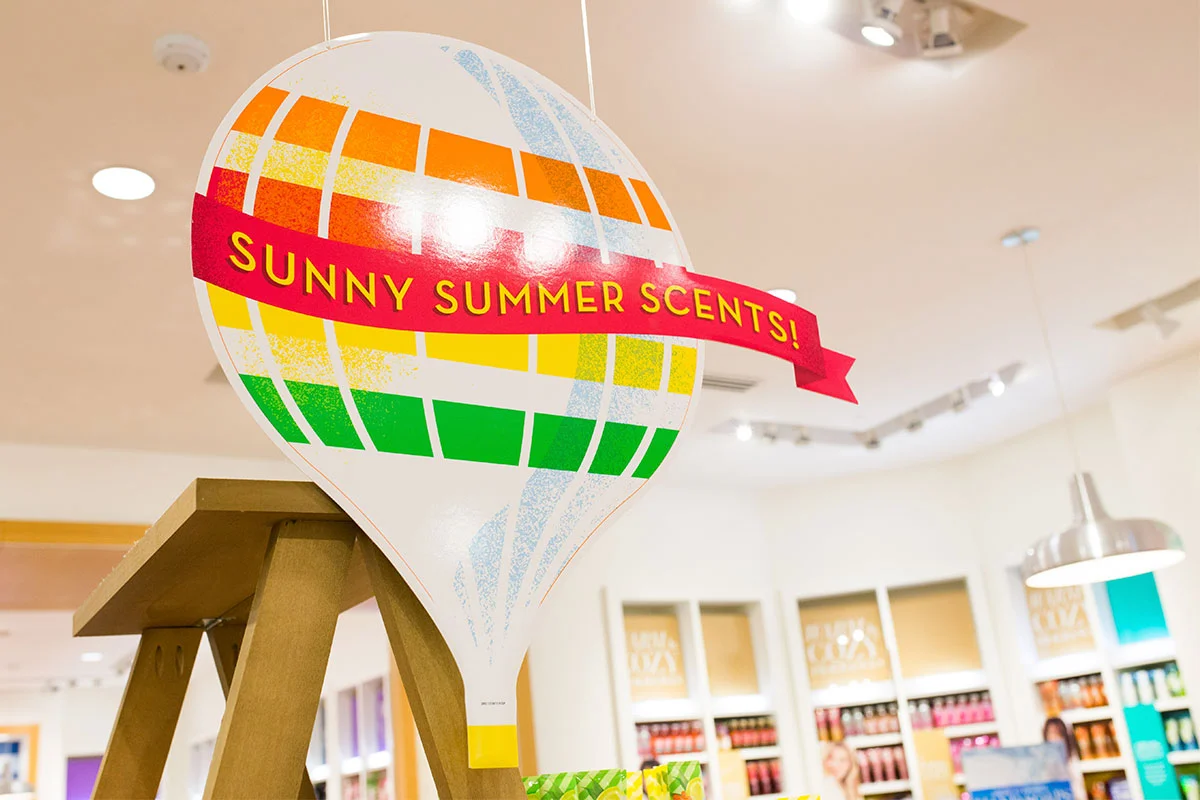 Colorful hot air balloon display with a banner reading "Sunny Summer Scents!" inside a store with shelves of scented products.