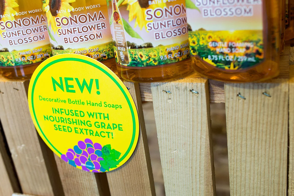 Display of Sonoma Sunflower Blossom hand soaps in bottles on a wooden shelf, with a bright yellow sign announcing new decorative bottle hand soaps infused with nourishing grape seed extract.