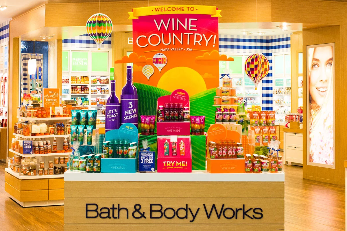 A Bath & Body Works store display promoting Vineyard Fresh fragrances with colorful bottles and packaging, a large welcome sign for 'Wine Country' featuring a sunset, hot air balloons, and rolling hills.