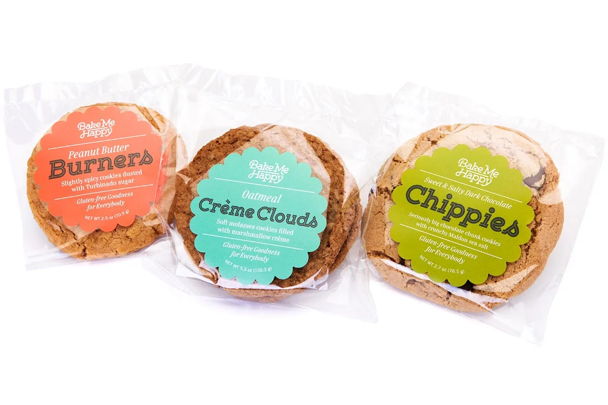Three packaged cookies from Bake Me Happy: Peanut Butter Burners, Creme Clouds oatmeal cookies, and Chippies dark chocolate chunk cookies, each in clear plastic wrapping with colorful labels.