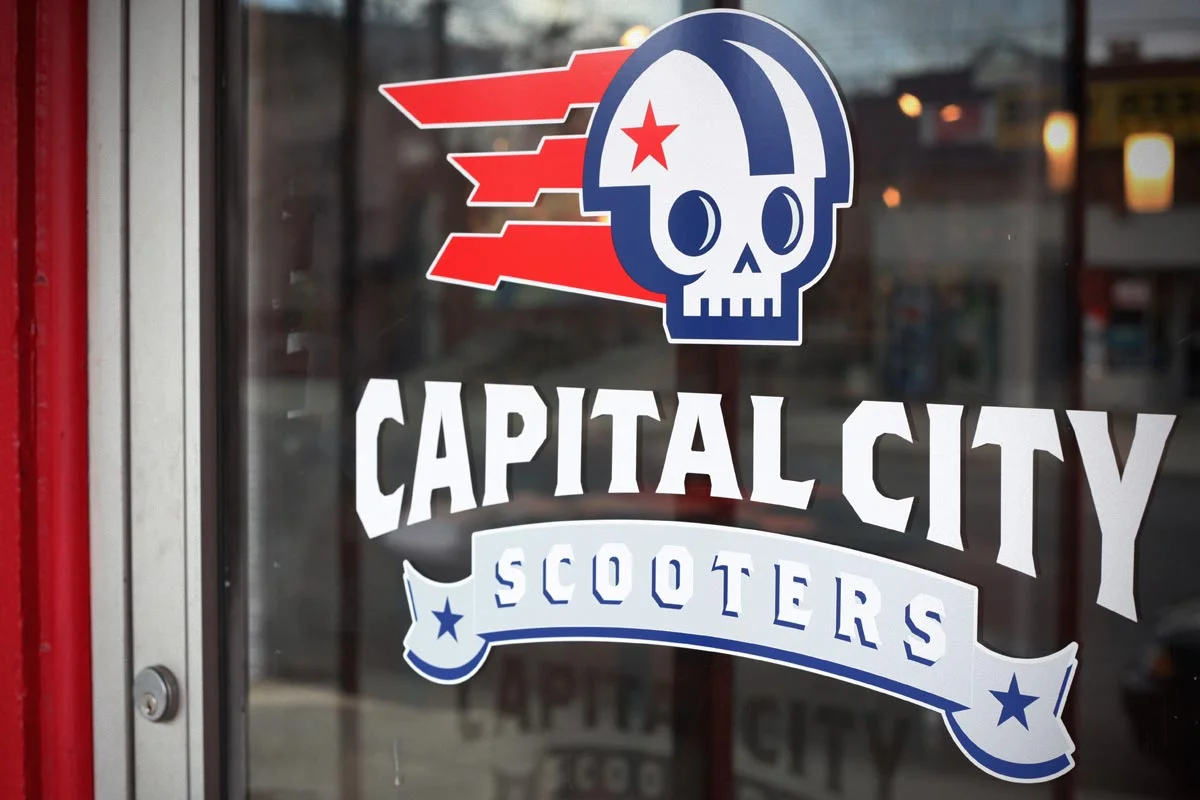 Sign for Capital City Scooters with a skull and helmet logo and red, white, and blue colors.