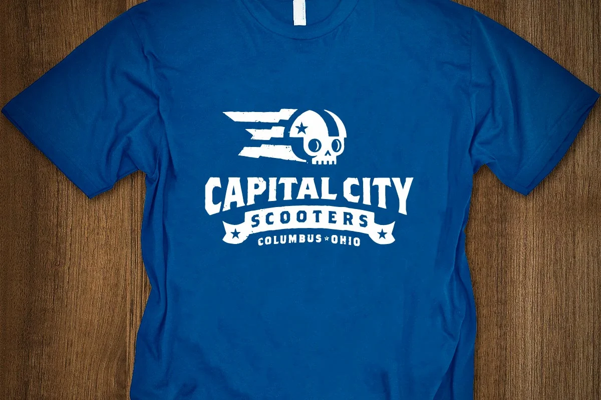Blue t-shirt with white graphic and text for Capital City Scooters in Columbus, Ohio, featuring a skull with a helmet and motion lines.