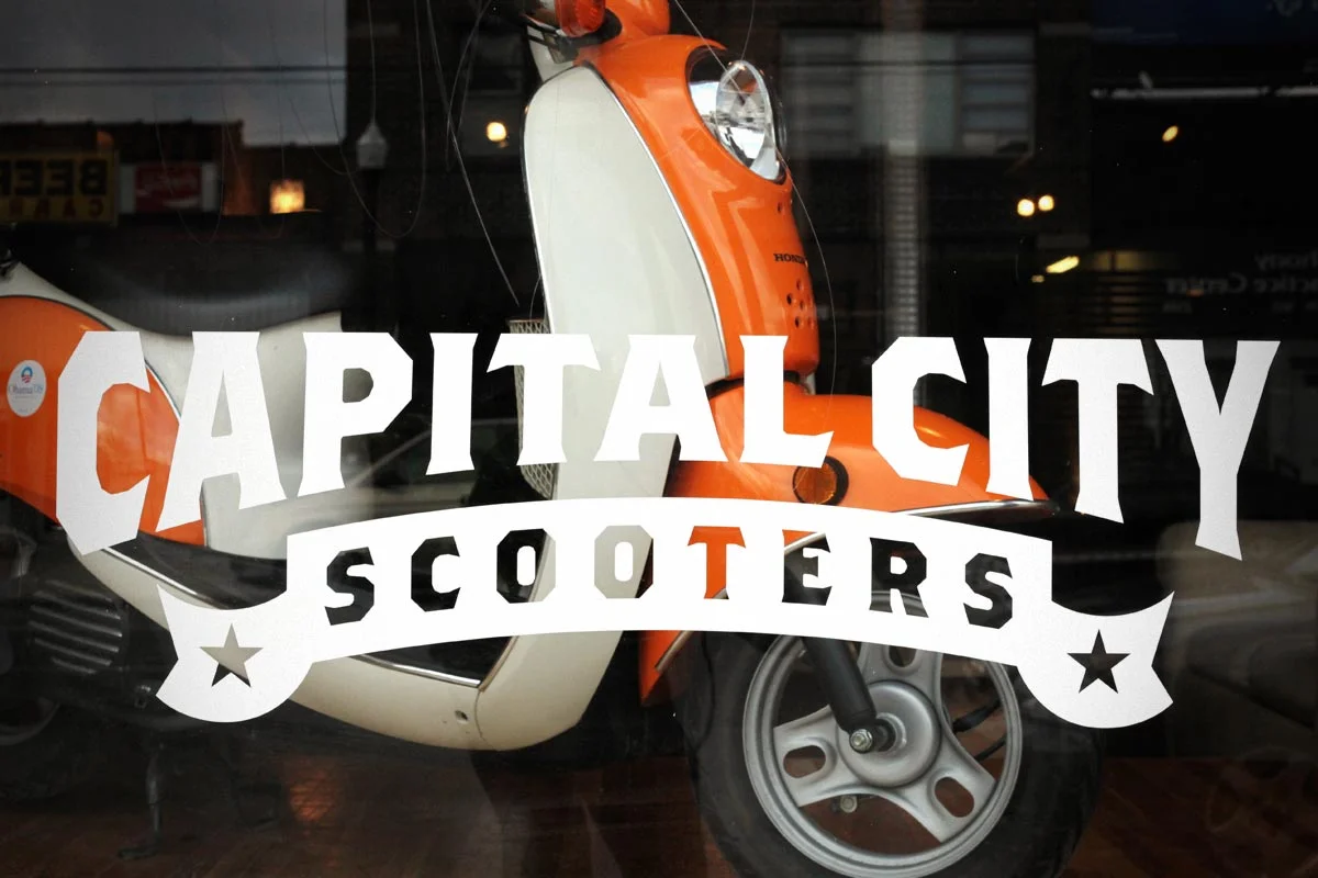 A storefront window with a sticker that reads "Capital City Scooters" in bold letters, featuring an illustration of an orange and white scooter behind the sticker.