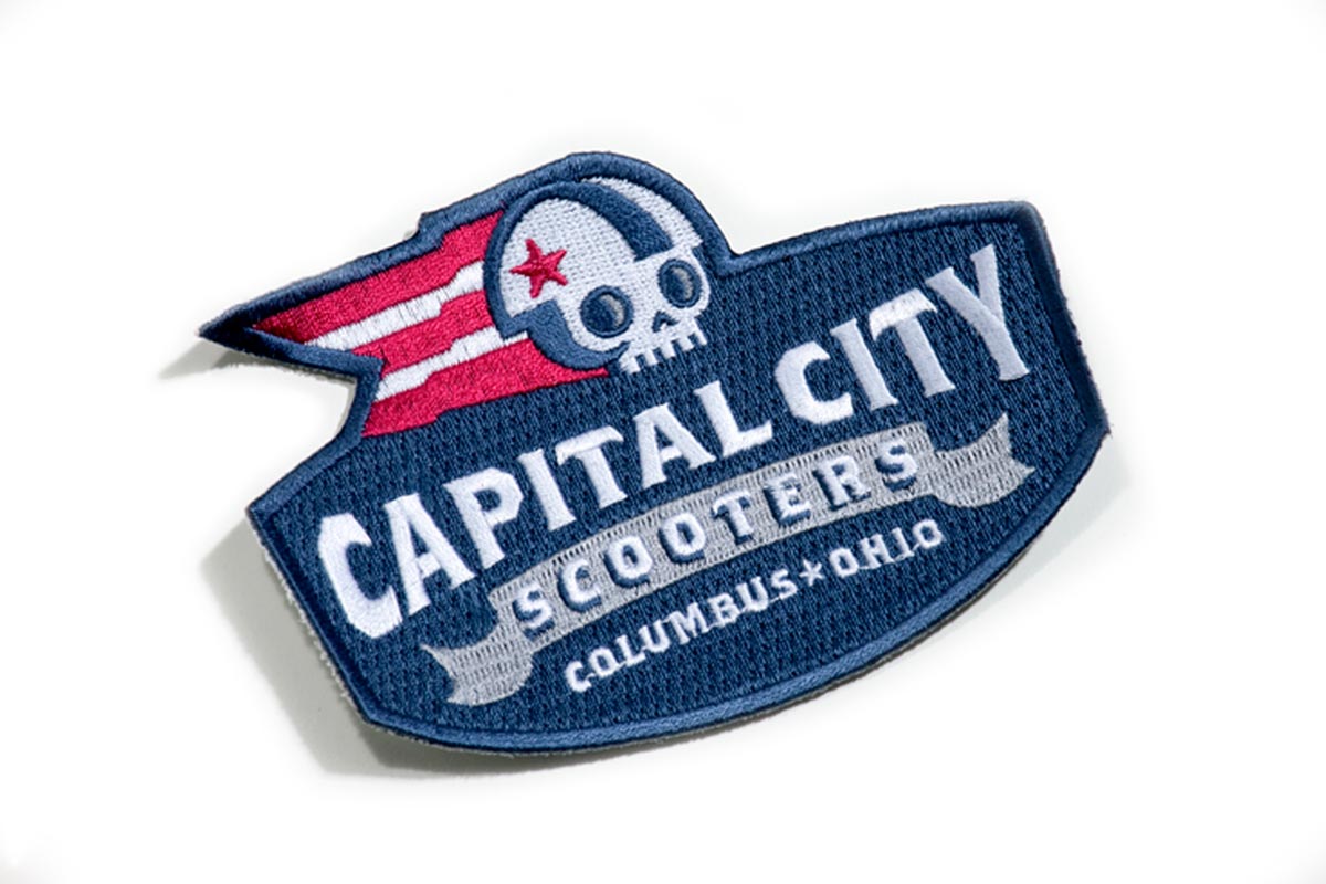 A patch with the logo of Columbus, Ohio, featuring a skull wearing headphones and a red, white, and blue color scheme, with the words "Capital City" and "Scooters".