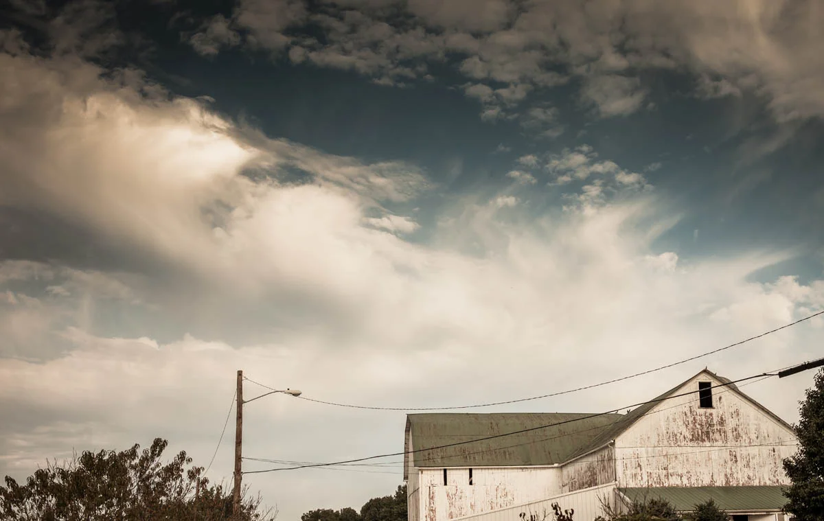 A weathered white barn with a green roof under a cloudy sky, with power lines and trees in the foreground.