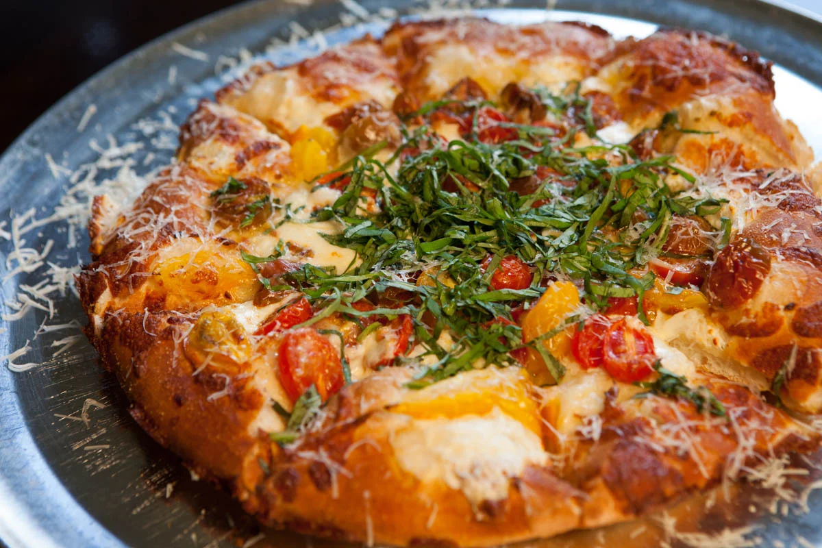 Close-up of a freshly baked pizza topped with cheese, cherry tomatoes, bell peppers, and fresh basil on a metal tray.