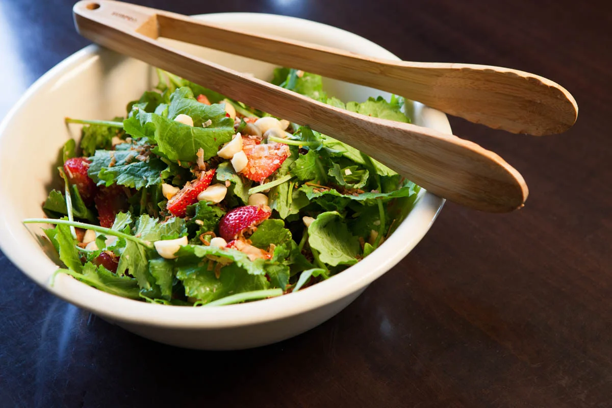 Bowl of fresh mixed salad with greens, strawberries, and nuts on dark wooden table, with wooden salad tongs across the top.