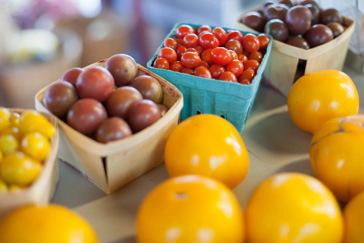 Assorted fresh produce including yellow tomatoes, red cherry tomatoes, grape tomatoes, and purple tomatoes displayed in baskets at a market.