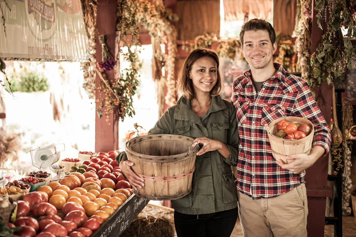 A smiling man and woman holding baskets of fresh tomatoes at a farmer's market, with a display of apples and other produce behind them.