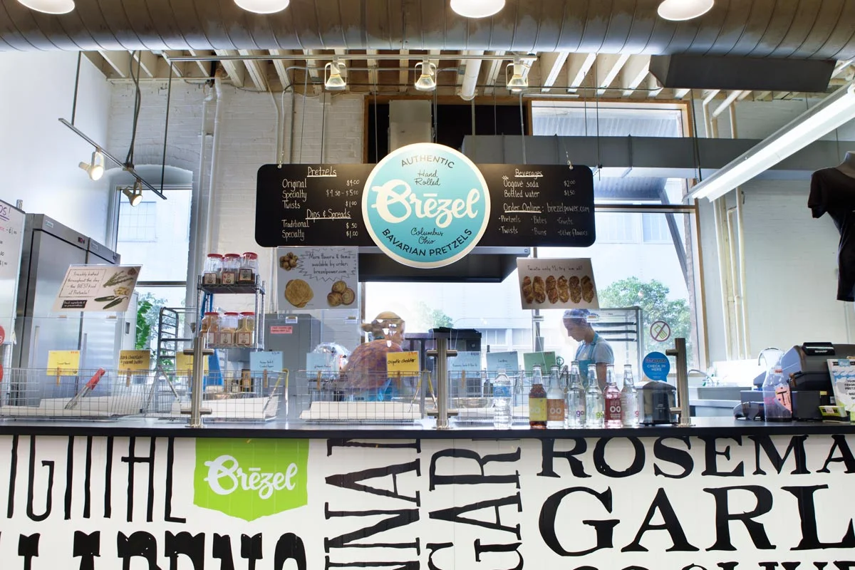 Counter at a pretzel shop called Ozzel, with a menu board and employees serving customers. Signage and menu items related to Bavarian pretzels and beverages are visible.
