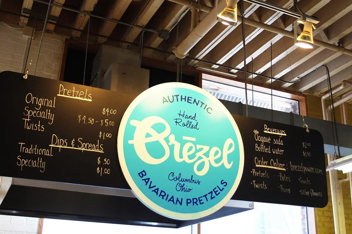 Signboard of Oriezl Bavarian Pretzels with menu prices for pretzels, dips, spreads, drinks, and online ordering, inside a bakery or cafe.