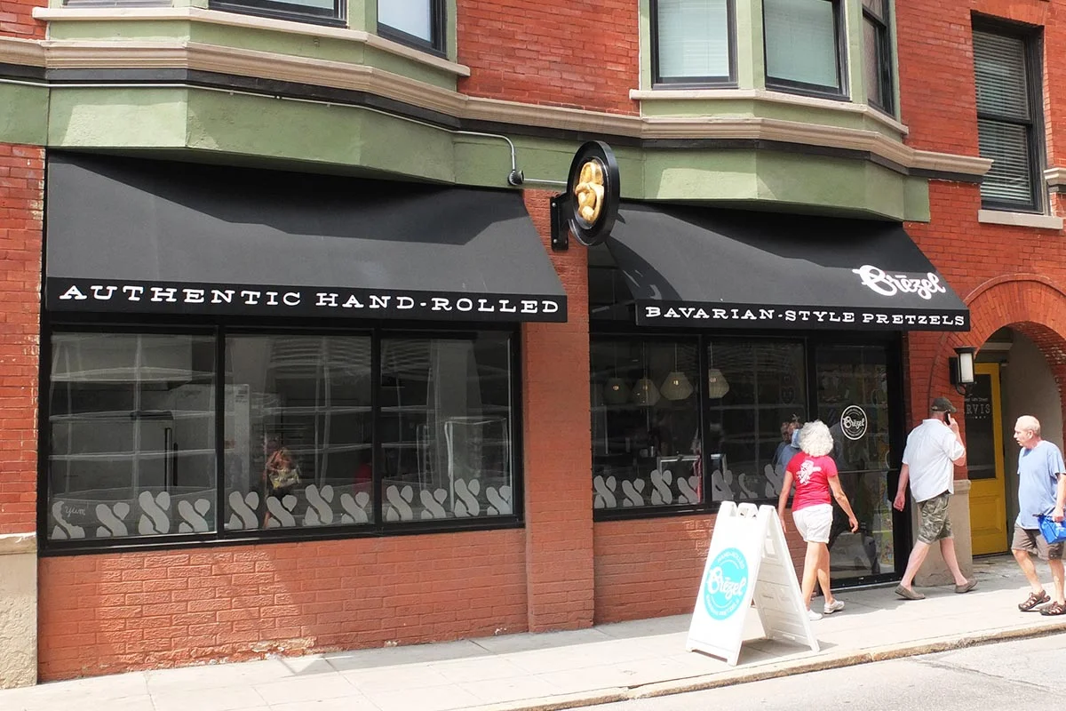 Storefront with black awnings advertising authentic hand-rolled Bavarian-style pretzels, with people walking on the sidewalk in front.