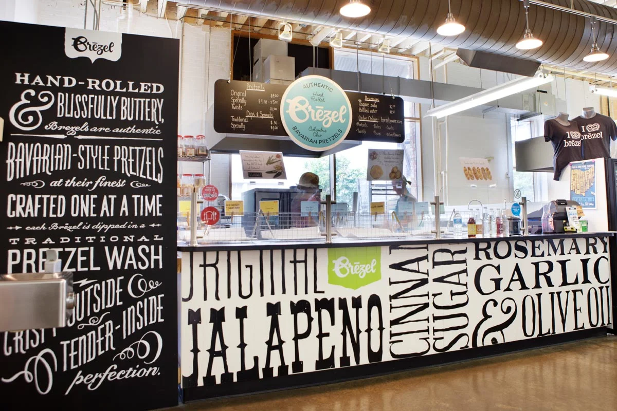 A counter at a bakery or pretzel shop with signage describing hand-rolled Bavarian pretzels, donuts, and traditional pretzel wash. The black and white decor features large, bold text with words like JALAPENO, CAYENNE, and GARLIC, and there are manneq